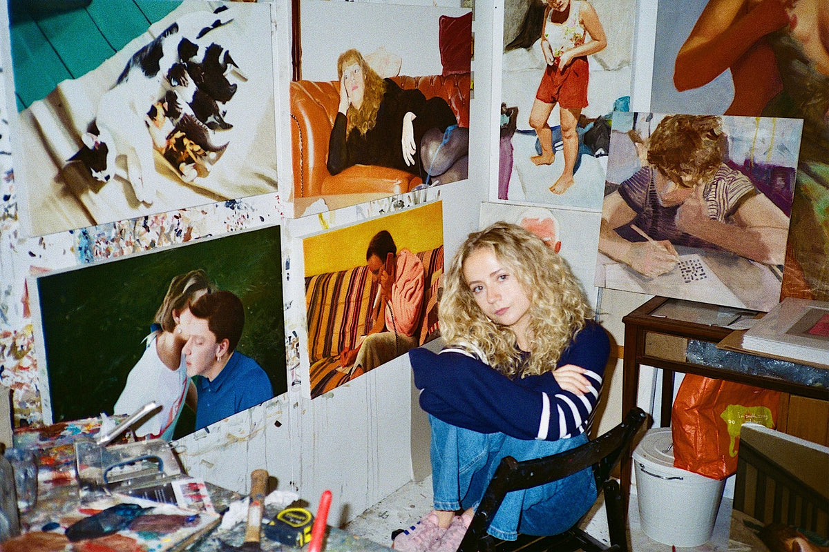 Artist Georgie Huxley with blonde curly hair sitting in her studio surrounded by figurative oil paintings for Partnership Editions.