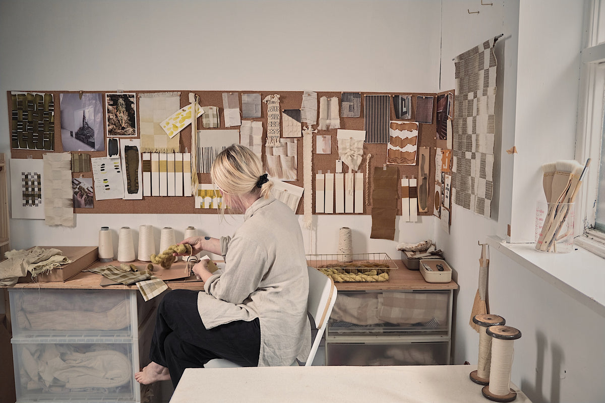 Textile artist Josie Mond-James working at her studio desk in front of a cork moodboard filled with fabric swatches and colour studies.