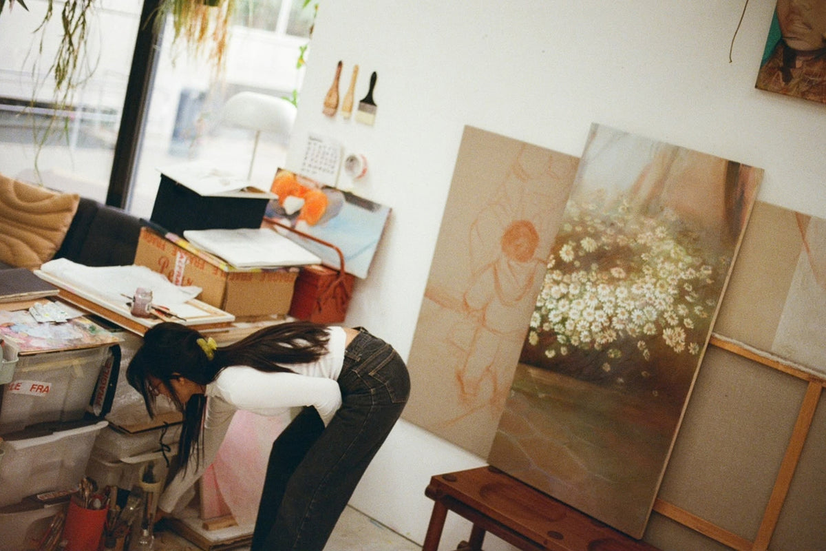 Artist Sophia Rosenthal bending down in her sunlit studio near large oil paintings on canvas and art supplies.