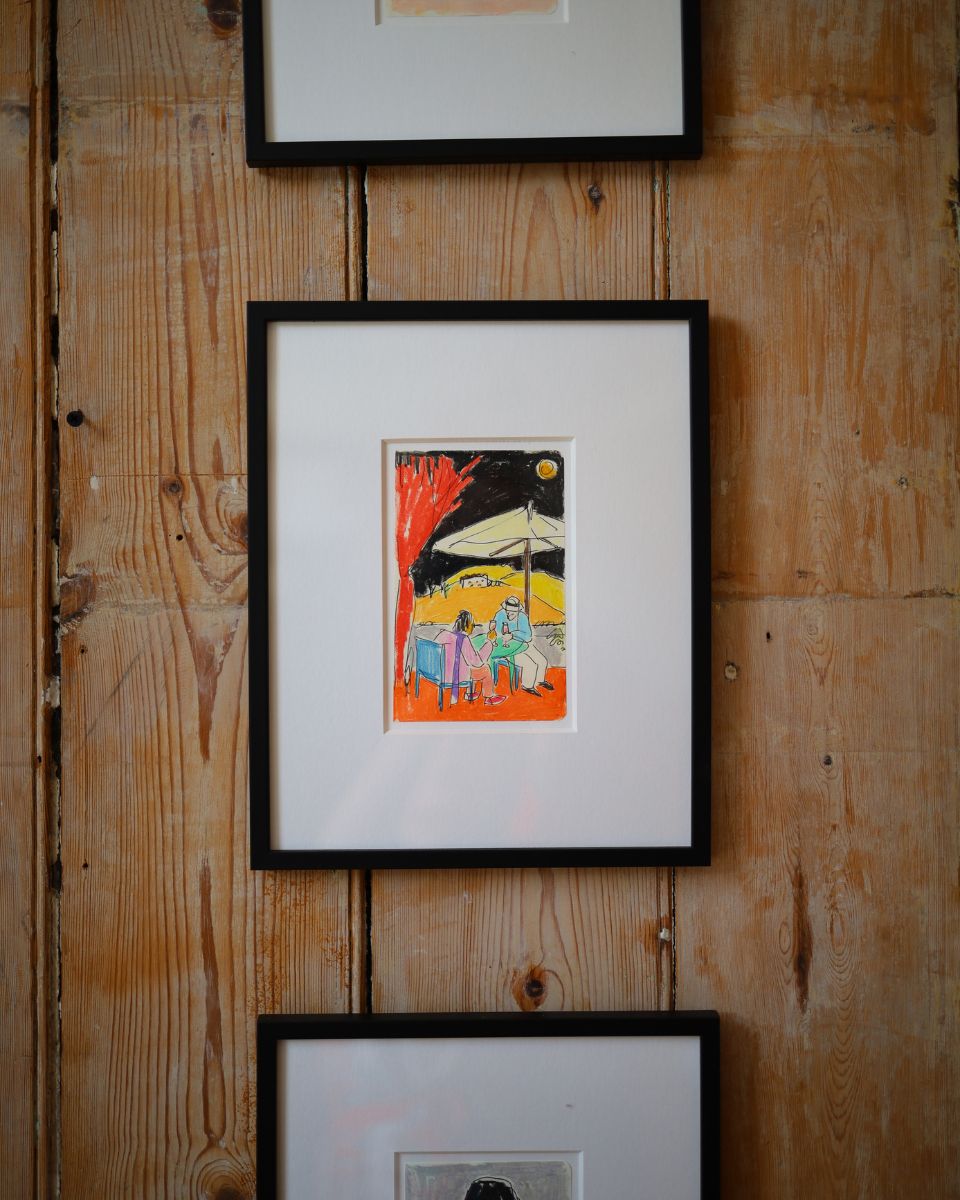 Close-up of a framed colourful illustration by Isabella Cotier depicting figures sitting under a cafe umbrella at night, hung on a rustic wooden wall.