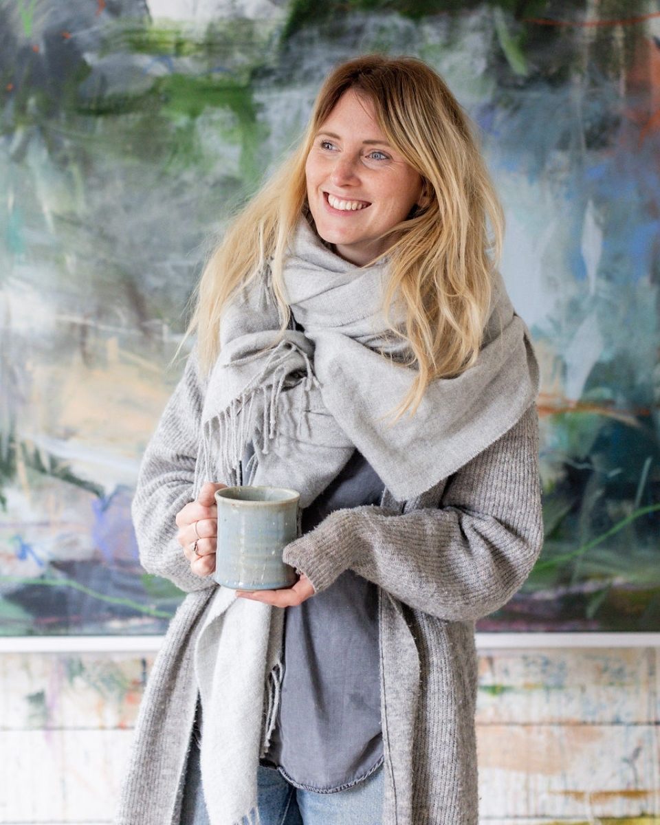 A portrait of a smiling Bethany Holmes relaxing inside her art studio while holding a cup of tea.