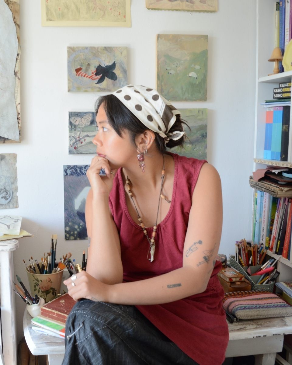 A reflective portrait of artist Bethenie Carriaga sat in front of detailed artworks and sketches studio wall.