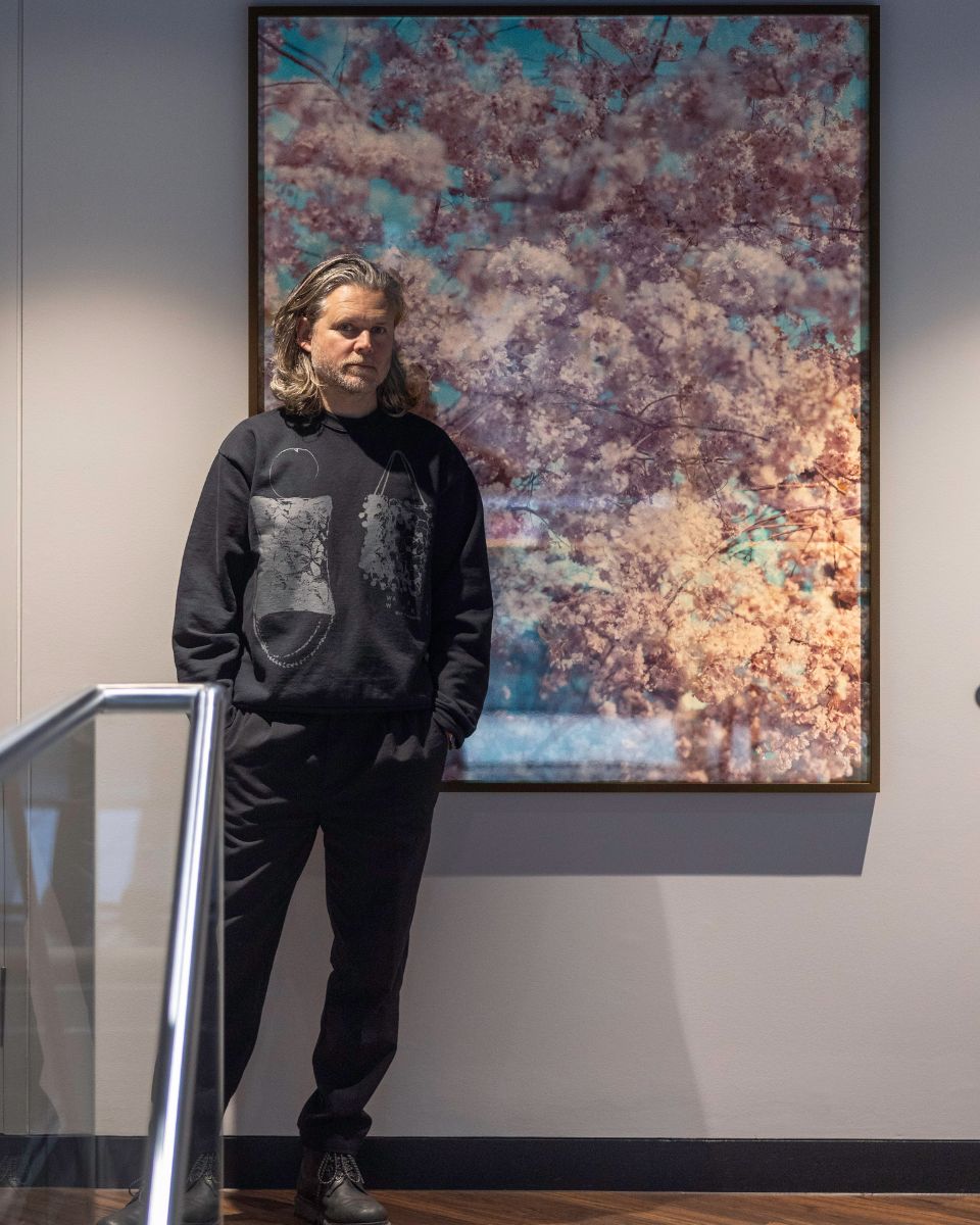 Artist Casey Moore standing beside a large framed photograph of pink cherry blossoms in a gallery setting.