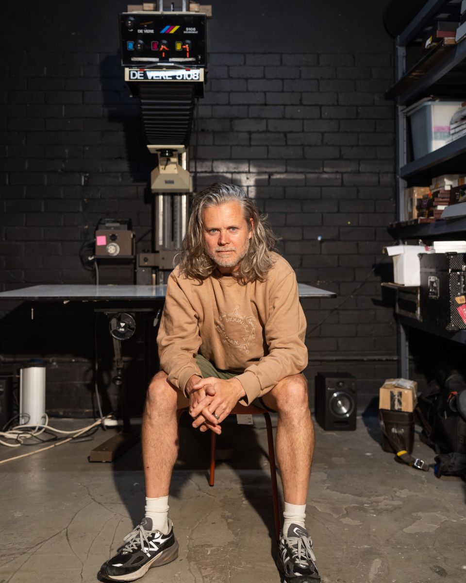 Photographer Casey Moore seated in his darkroom studio in front of a De Vere 5108 enlarger machine.