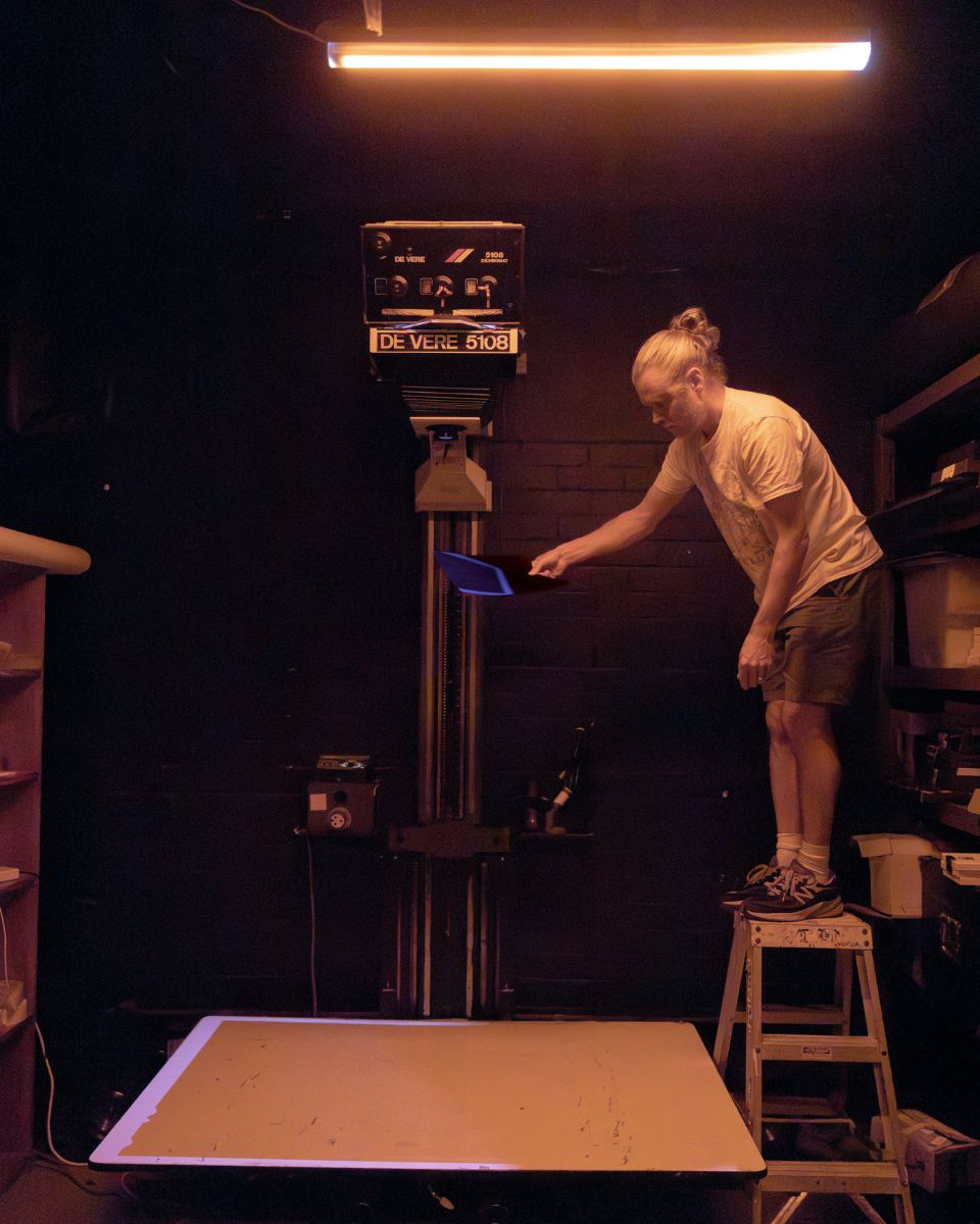 Casey Moore standing on a step stool in a darkroom to adjust exposure under a photographic enlarger.