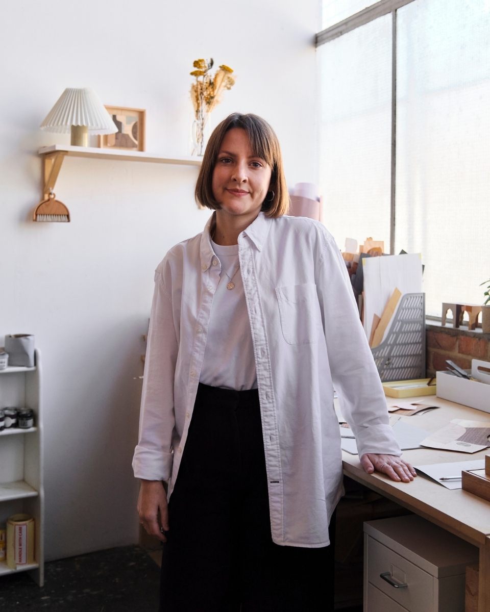 Artist Chelsea Vivash standing by her studio desk surrounded by wooden shelves and framed marquetry artworks.