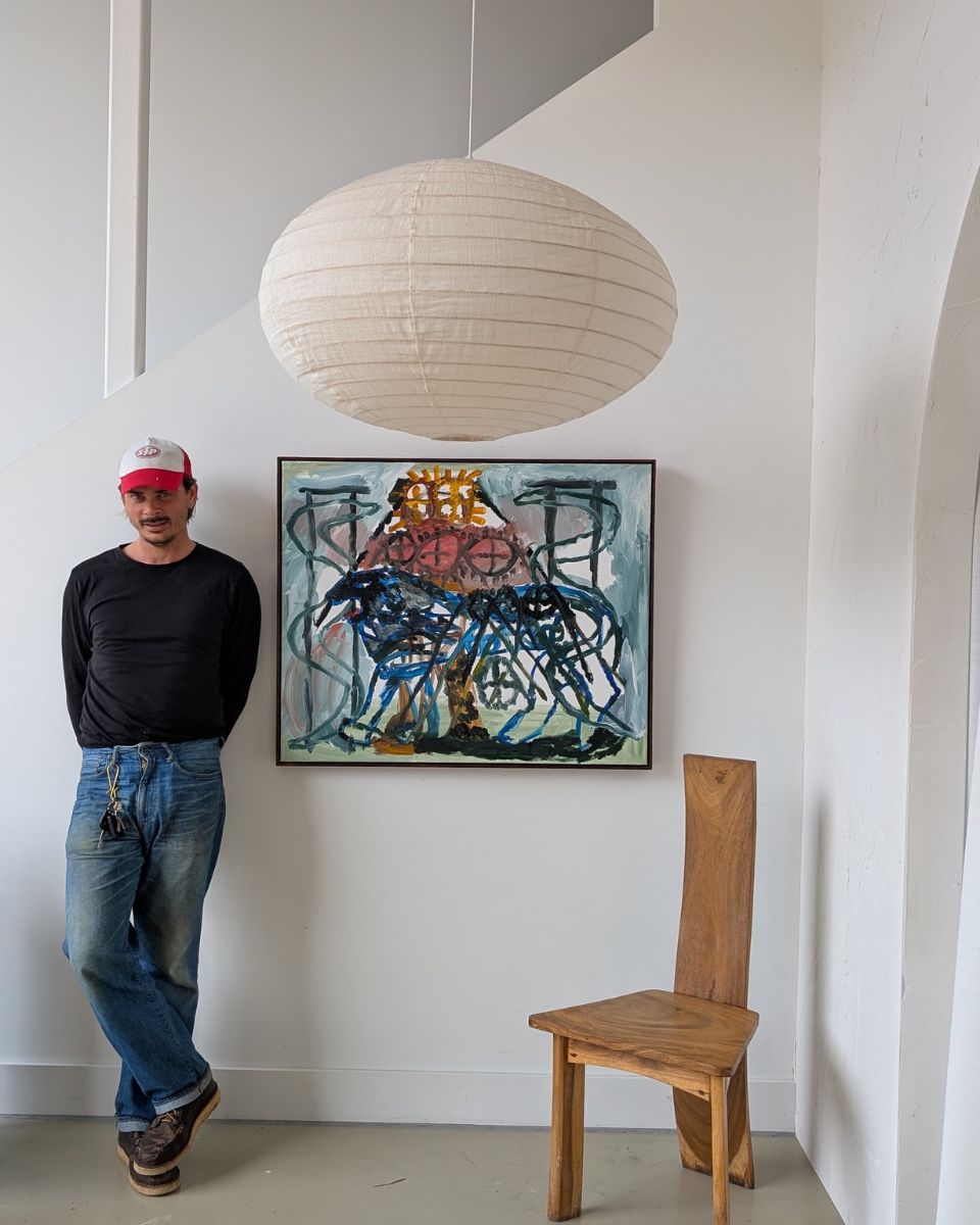 Portrait of contemporary artist Darcy McCrae standing beside a large framed figurative painting.