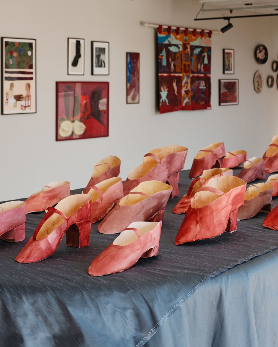 A detailed close-up shot of Phoebe Howard's paper shoes displayed on a table in front of framed, mounted artworks by contemporary artists at the Partnership Editions 'Dressing Up' exhibition.