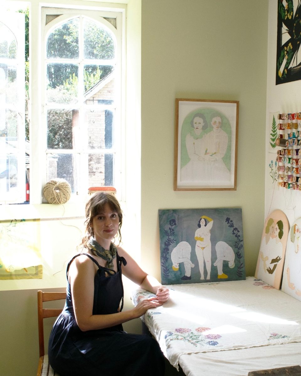 Artist Elizabeth Loveday at her home studio desk surrounded by colorful threads and framed artworks.