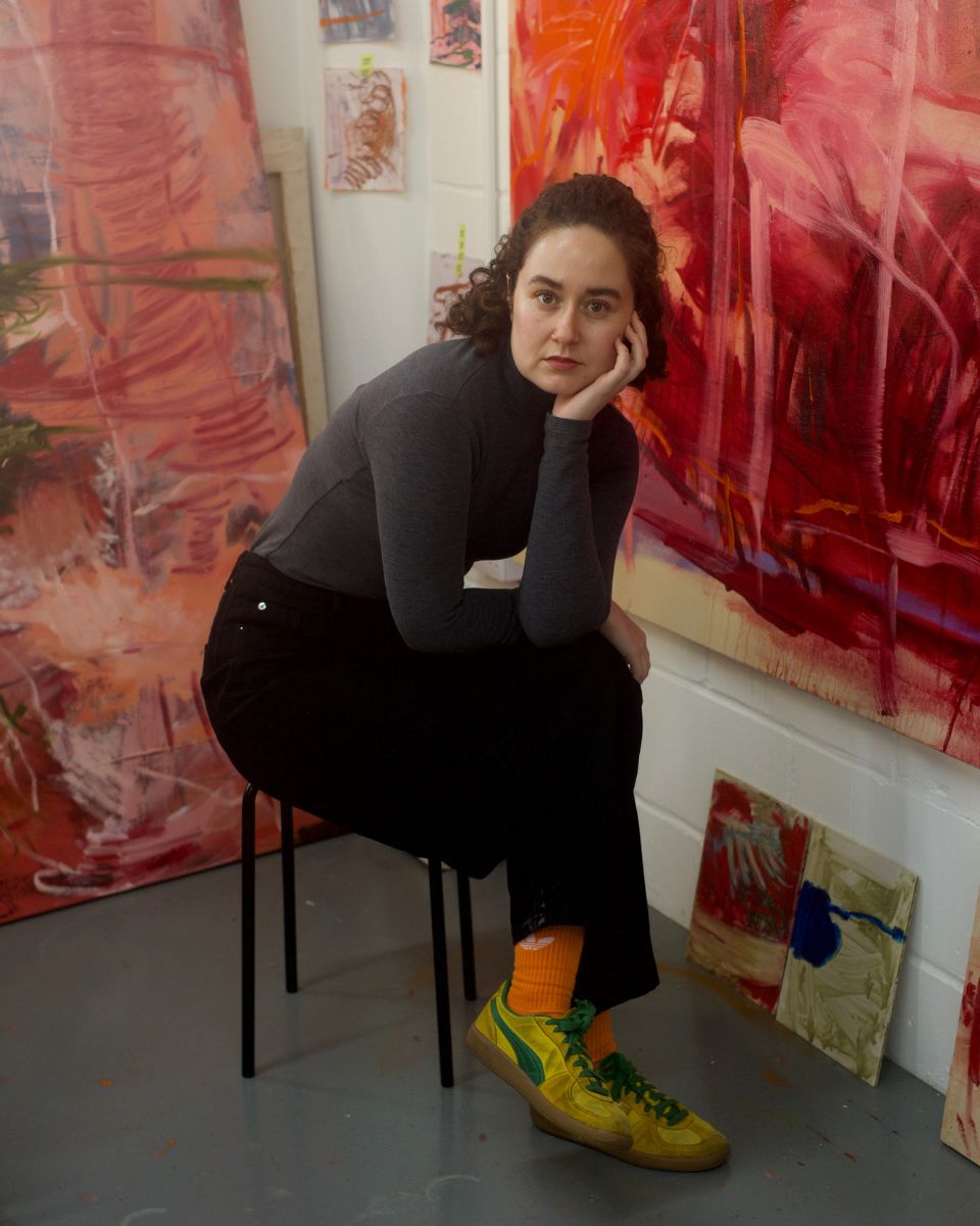Artist Ellie Walker seated in her studio surrounded by large pink and red abstract canvas paintings for Partnership Editions.