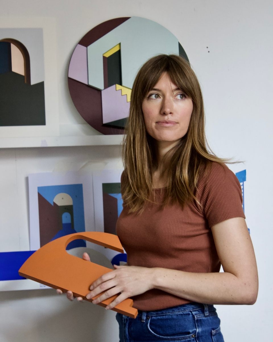 Artist Emily Forgot posing in her studio in front of a colorful, geometric architectural wall assemblage.