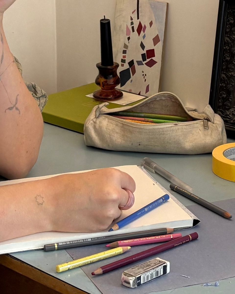 Close-up view of artist hands with coloured pencils, sketchbook, and eraser on a studio desk.