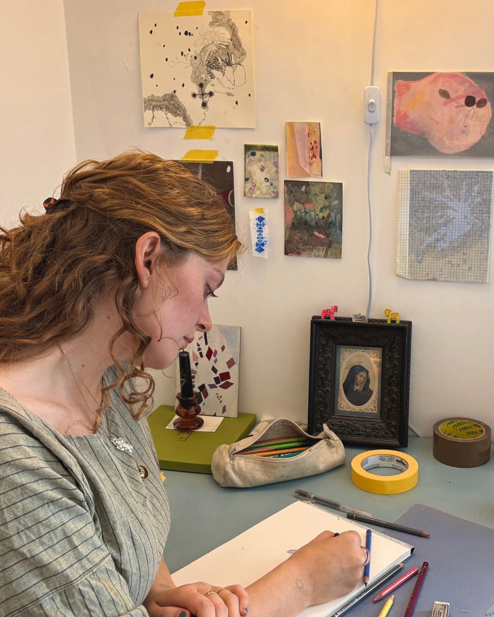 Artist Esme McMillan-Scott drawing with coloured pencils at her desk with sketches pinned to the wall.