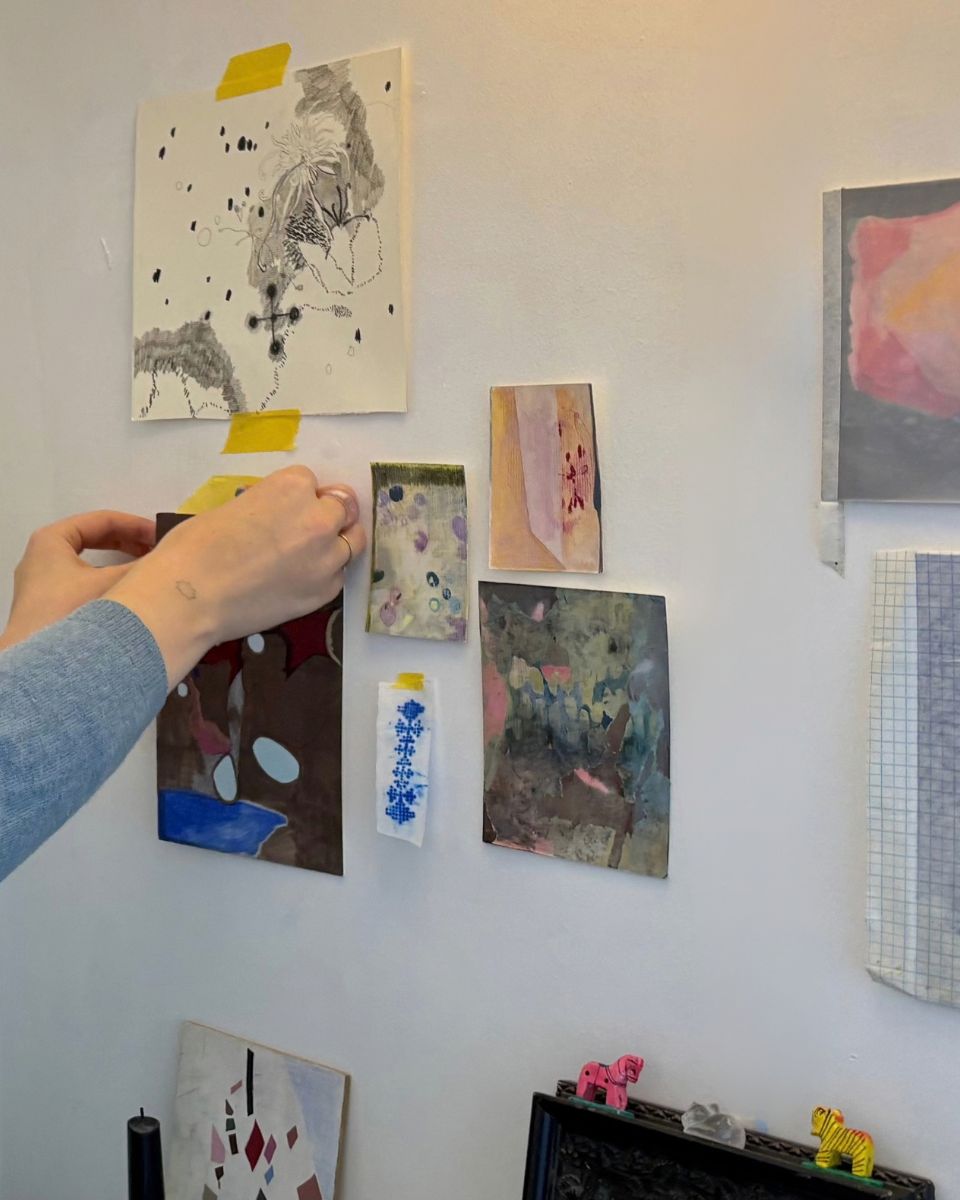 Artist hands taping small colourful paper works and mixed media studies to a white studio wall.