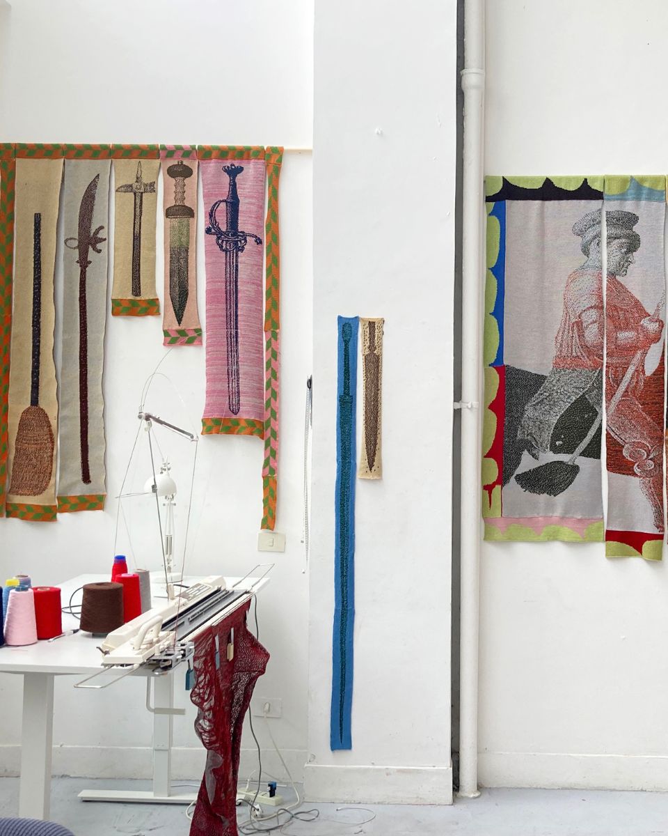 Knitting machine and colourful yarn spools on a desk in Eva Sajovic's studio with knitted sword textile art hanging on the wall.