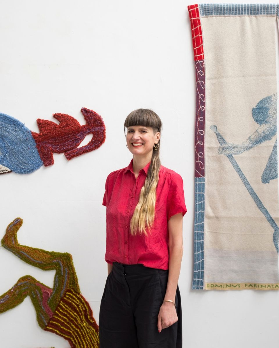 Portrait of artist Eva Sajovic wearing a red shirt, standing in front of her woven textile artworks in her studio.