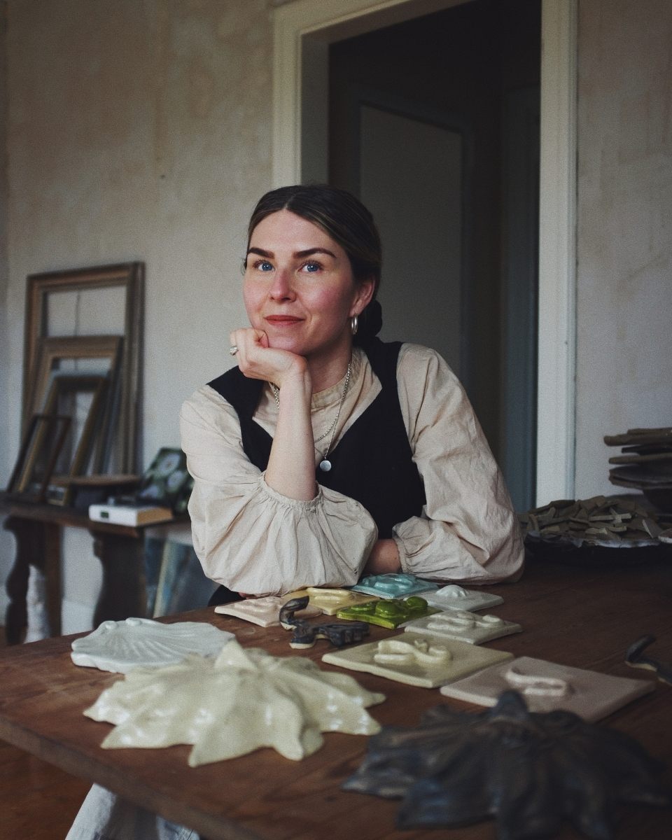 Ceramicist Fanny Schultz sitting at her studio desk in front of a collection of her handmade stoneware tiles.