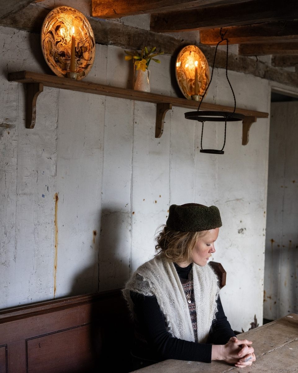 A moody portrait of artist Freyja Lee seated beneath a wooden shelf displaying two handmade ceramic sconces.