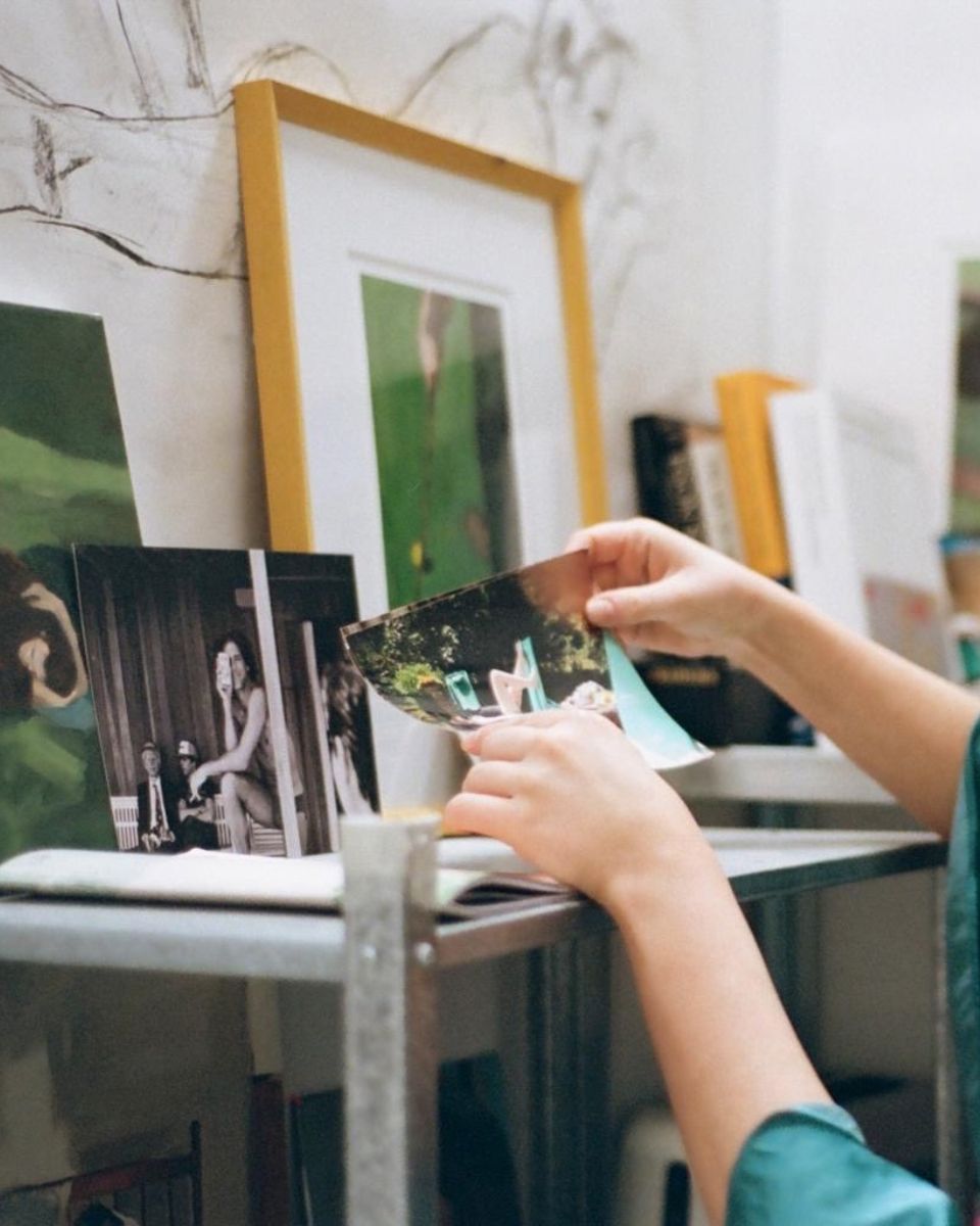 Artist hands holding reference photographs on a studio shelf with framed art in the background.