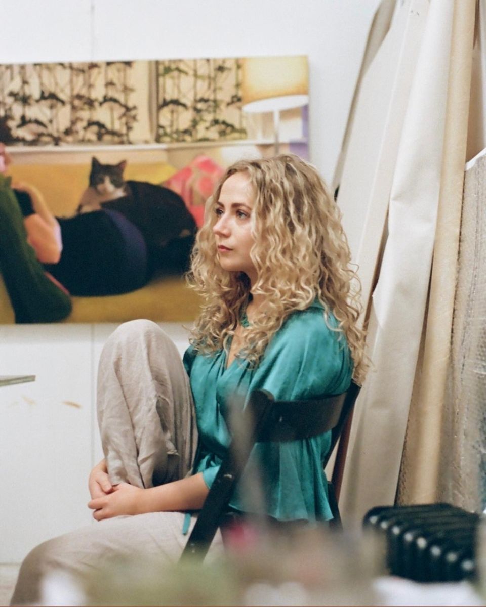 Portrait of artist Georgie Huxley in a teal blouse sitting in her studio beside a large figurative canvas.