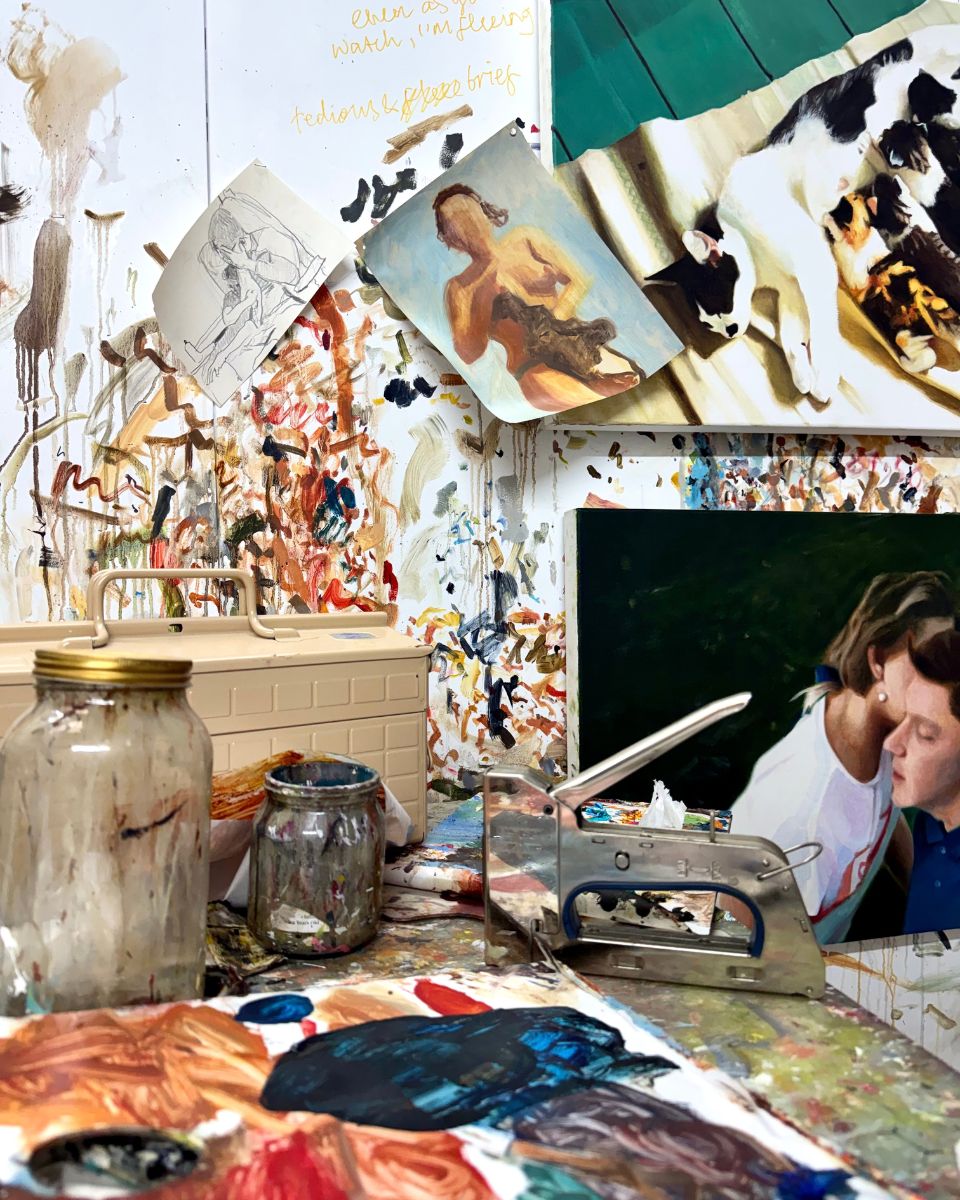 Artist studio desk with paint jars, staple gun, and reference sketches against a wall covered in paint splatters and handwritten notes.