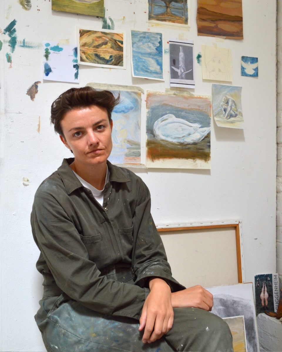 Portrait of artist Hattie Landells sitting in her studio in front of a gallery wall featuring work-in-progress drawings and small-scale paintings for Partnership Editions.