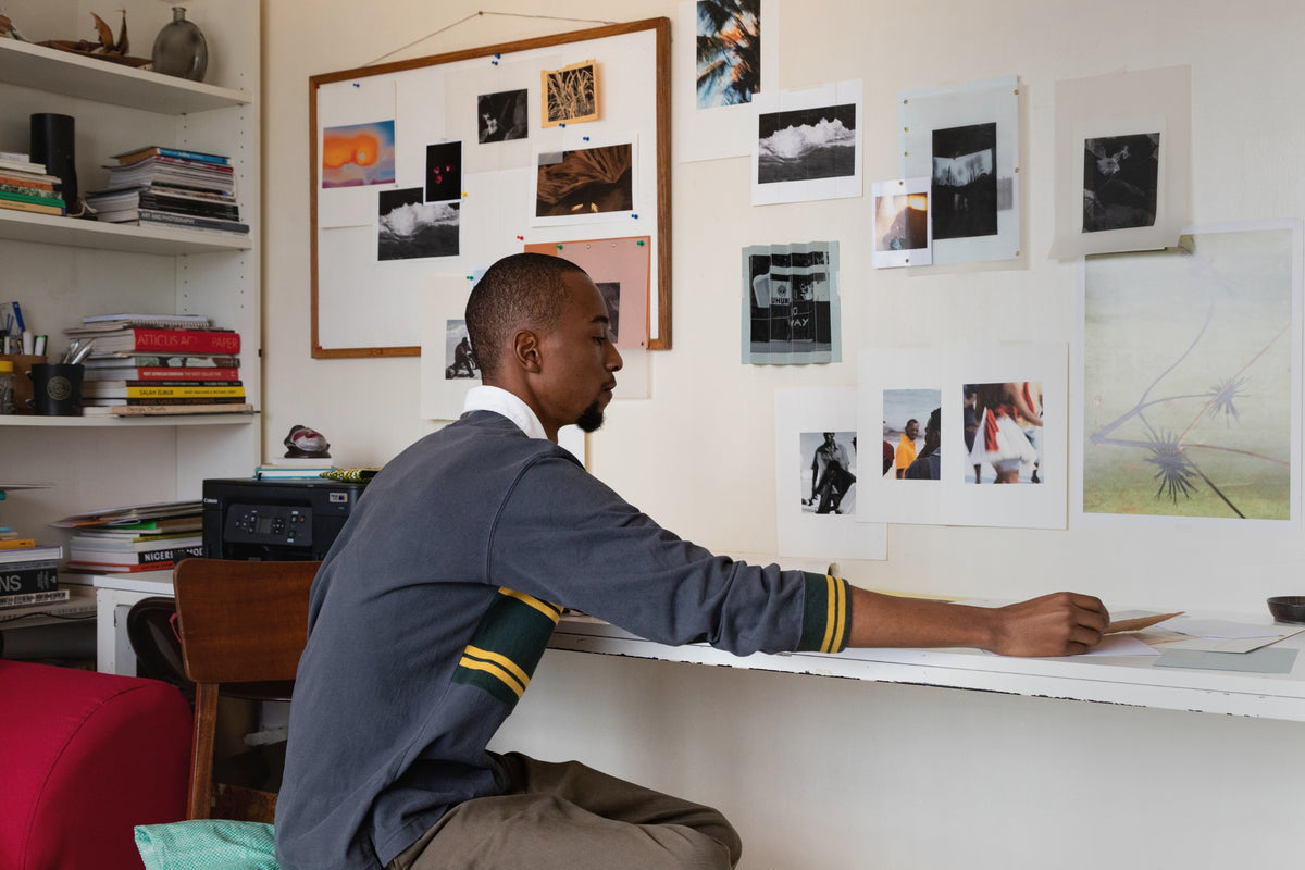 Photographer Ian Gichohi sitting at his studio desk examining prints, surrounded by mood boards and photograph displays for Partnership Editions.