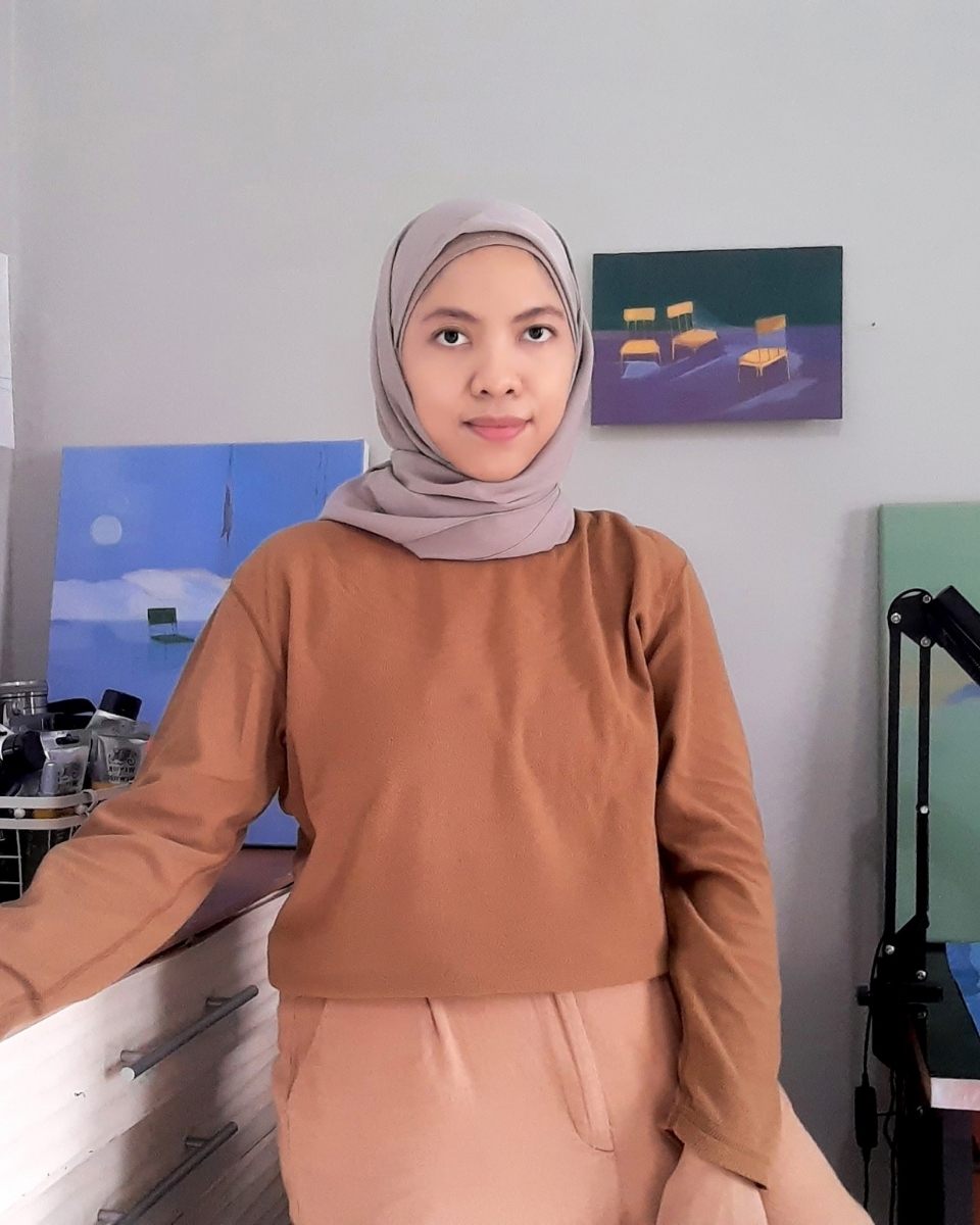 Artist Ifada Nisa posing in her studio in front of her dreamlike landscape paintings.