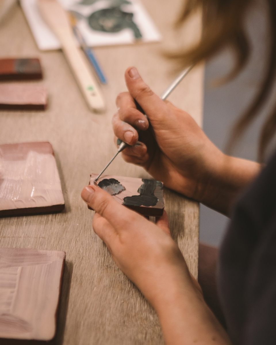 Close-up of Imogen Richards’ hands hand-painting terracotta tiles, showcasing the artisanal detail for Partnership Editions.