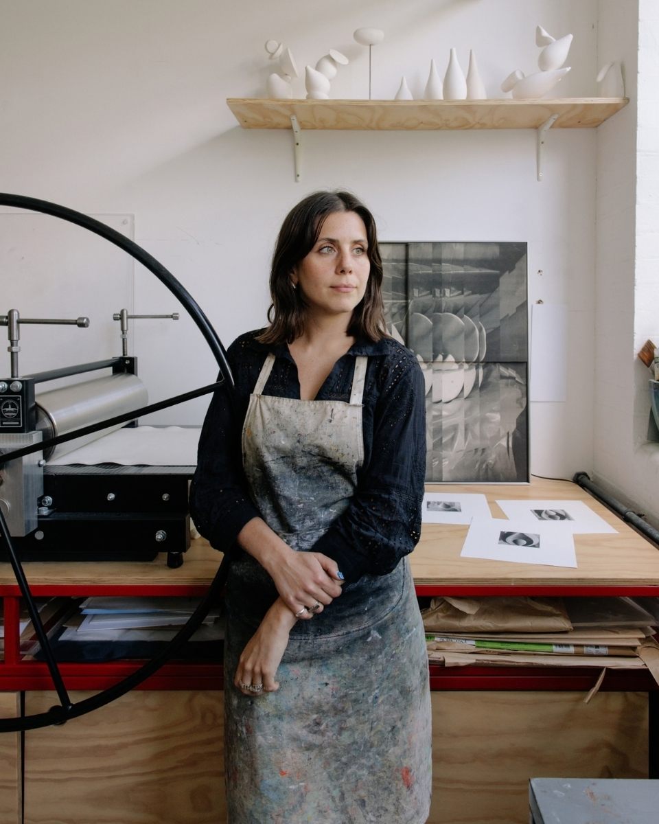 Artist Ines Fernandez de Cordova posing by the large metal spinning wheel of an etching press in her studio.