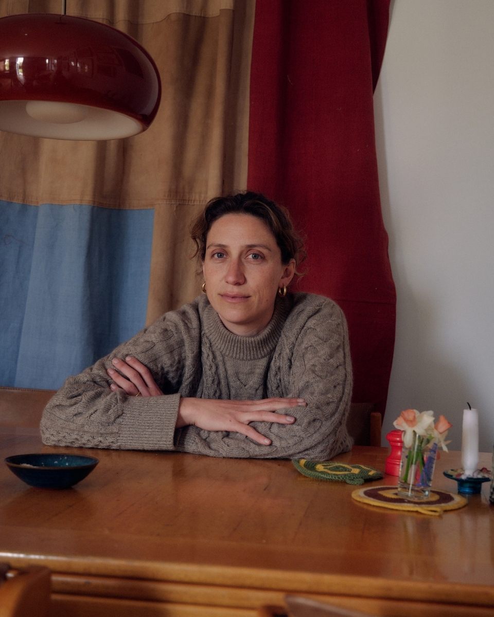 Textile artist Isabel Farchy posing at her studio table in front of a handcrafted linen patchwork curtain.