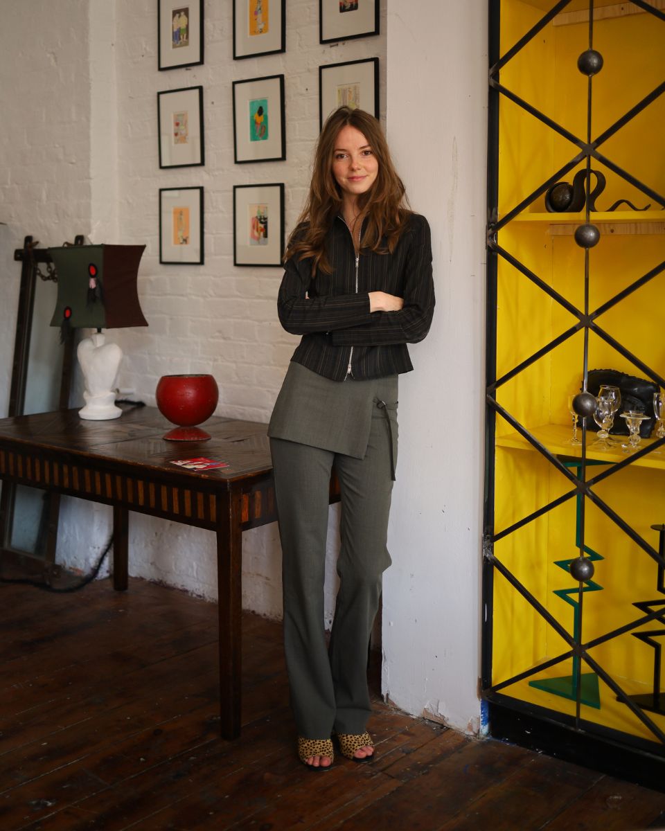 Portrait of artist Isabella Cotier standing with crossed arms in front of her artwork at the Cafe Culture exhibition.