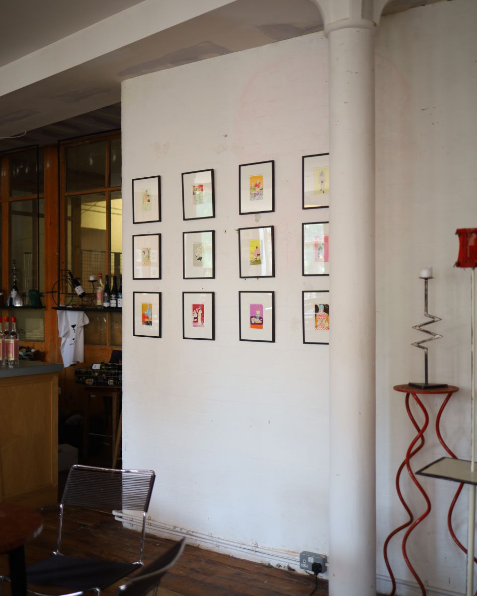 A grid display of twelve small, black-framed original illustrations by Isabella Cotier on a white brick gallery wall at the Cafe Culture exhibition.
