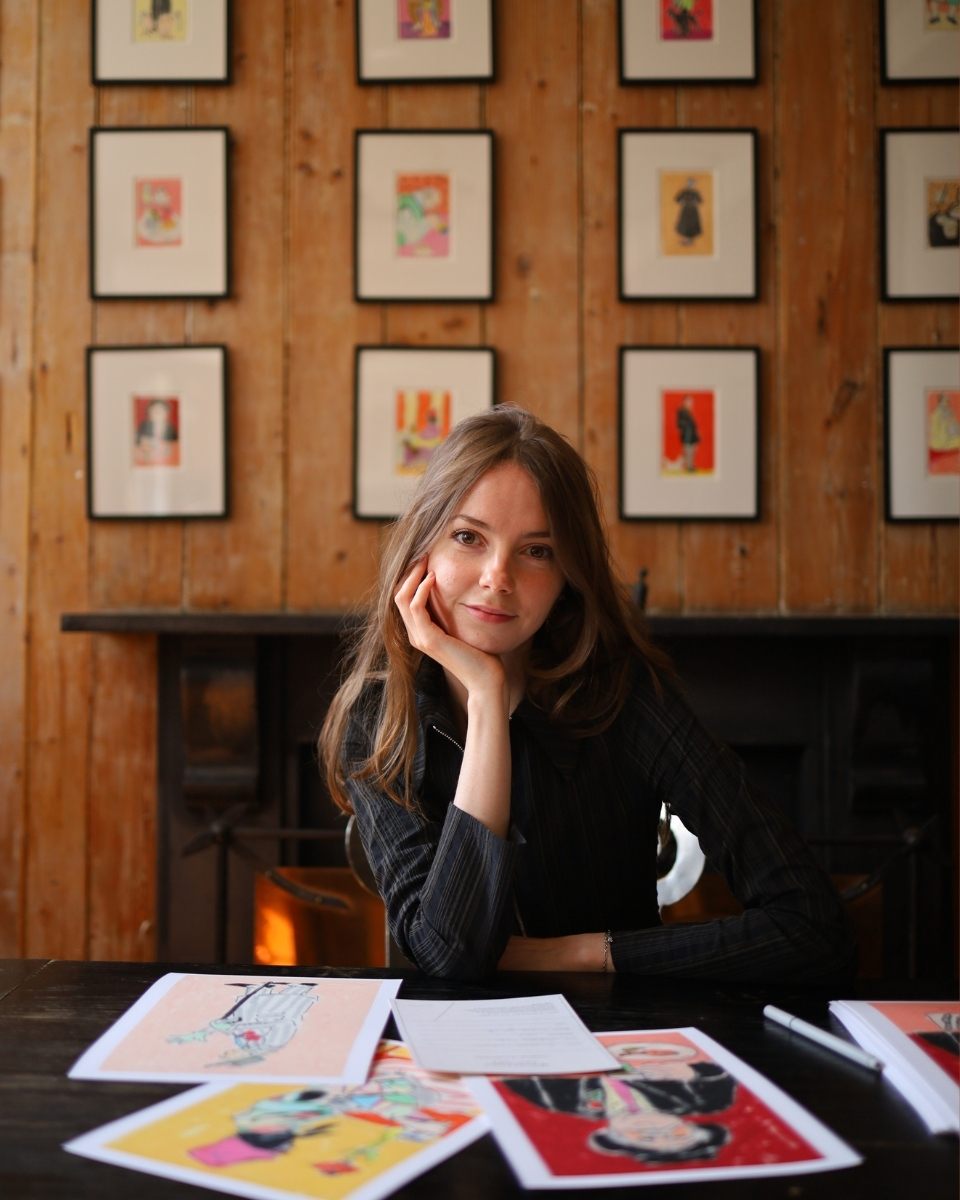 Isabella Cotier sitting at her signing table in front of her framed artworks at the Riverside Cafe.