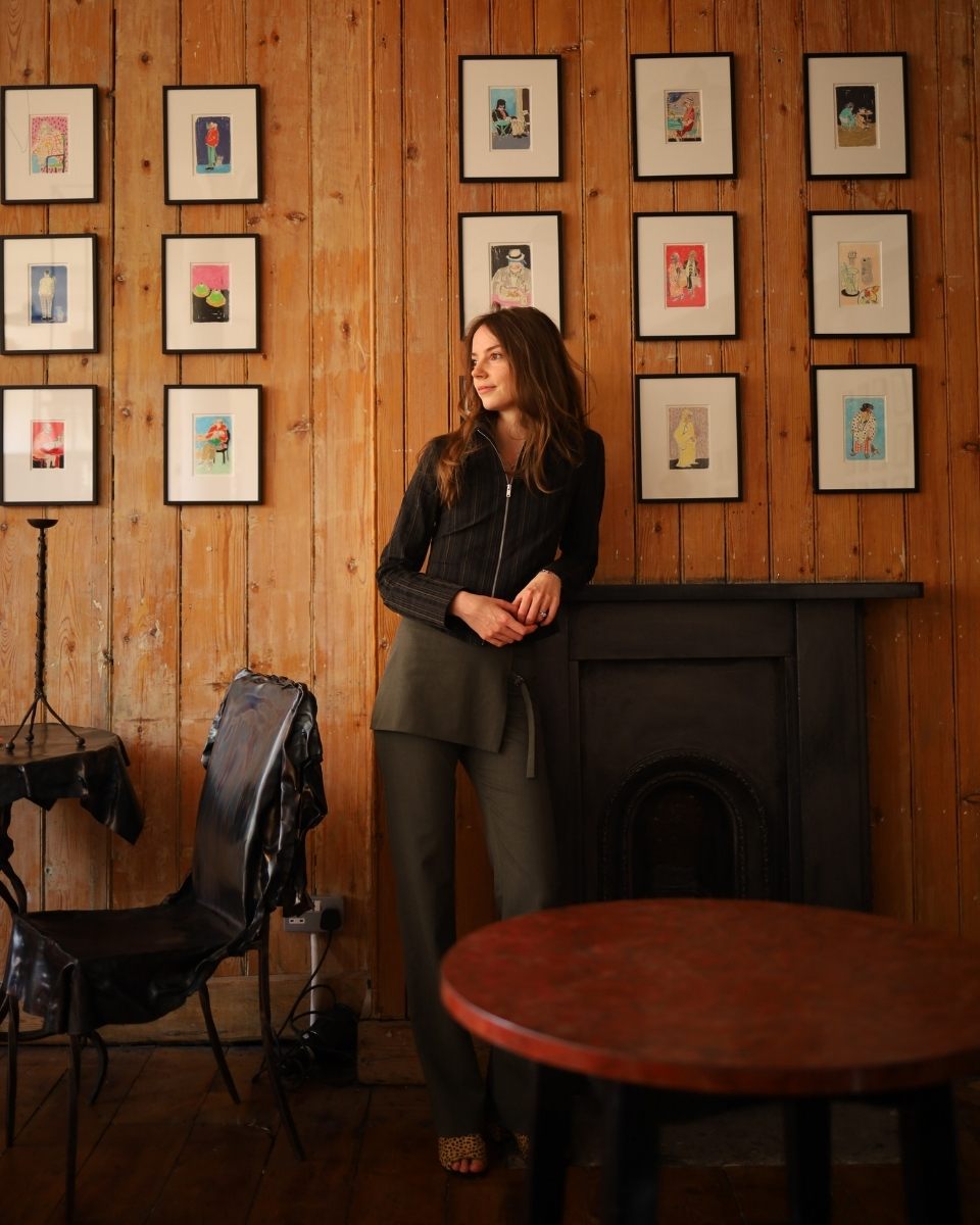 Portrait of artist Isabella Cotier at New River Cafe for her Cafe Culture exhibition, standing in front of a wall showcasing her framed figurative prints.
