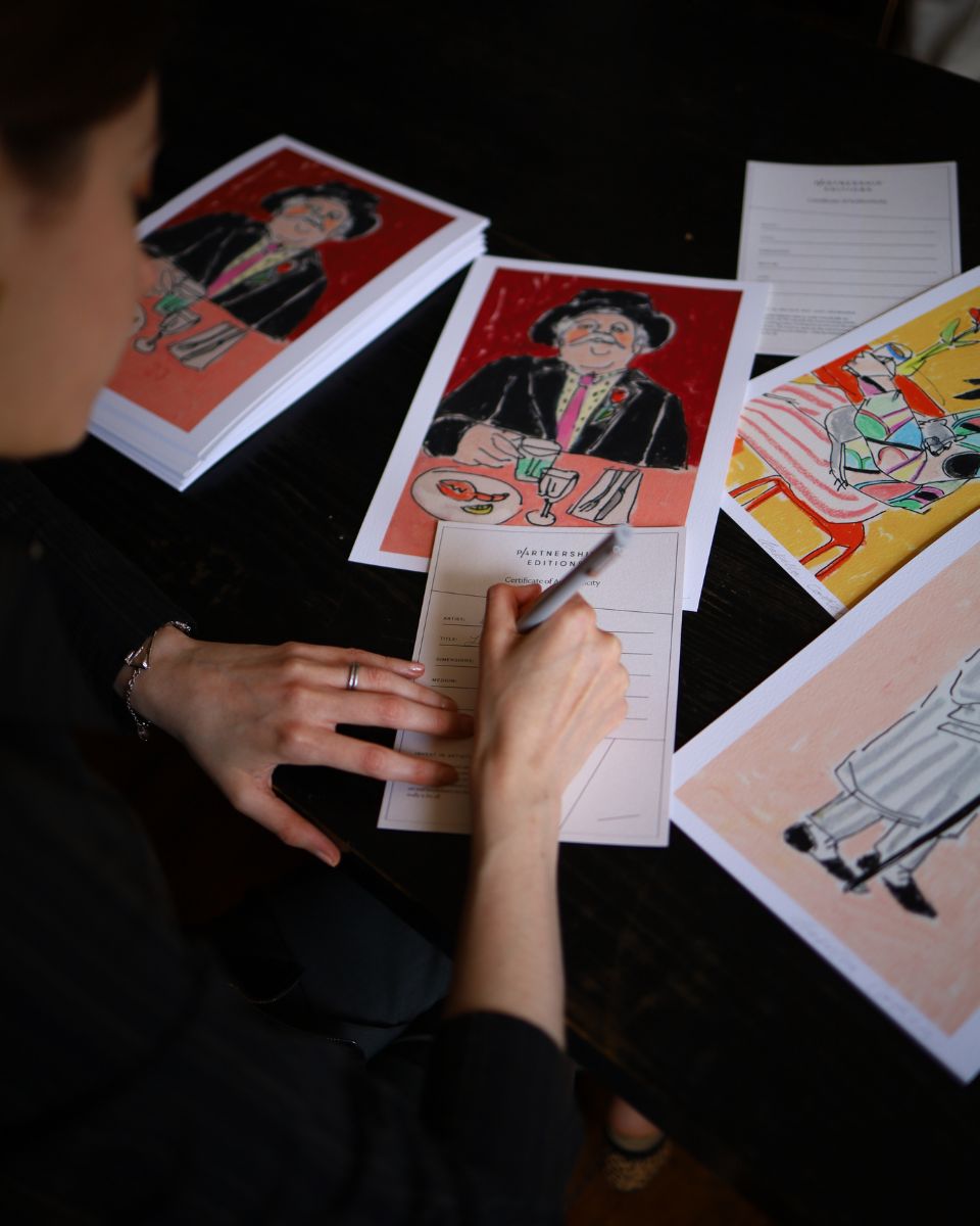 Over-the-shoulder view of artist Isabella Cotier filling out Partnership Editions certificates of authenticity next to her original cafe art prints.