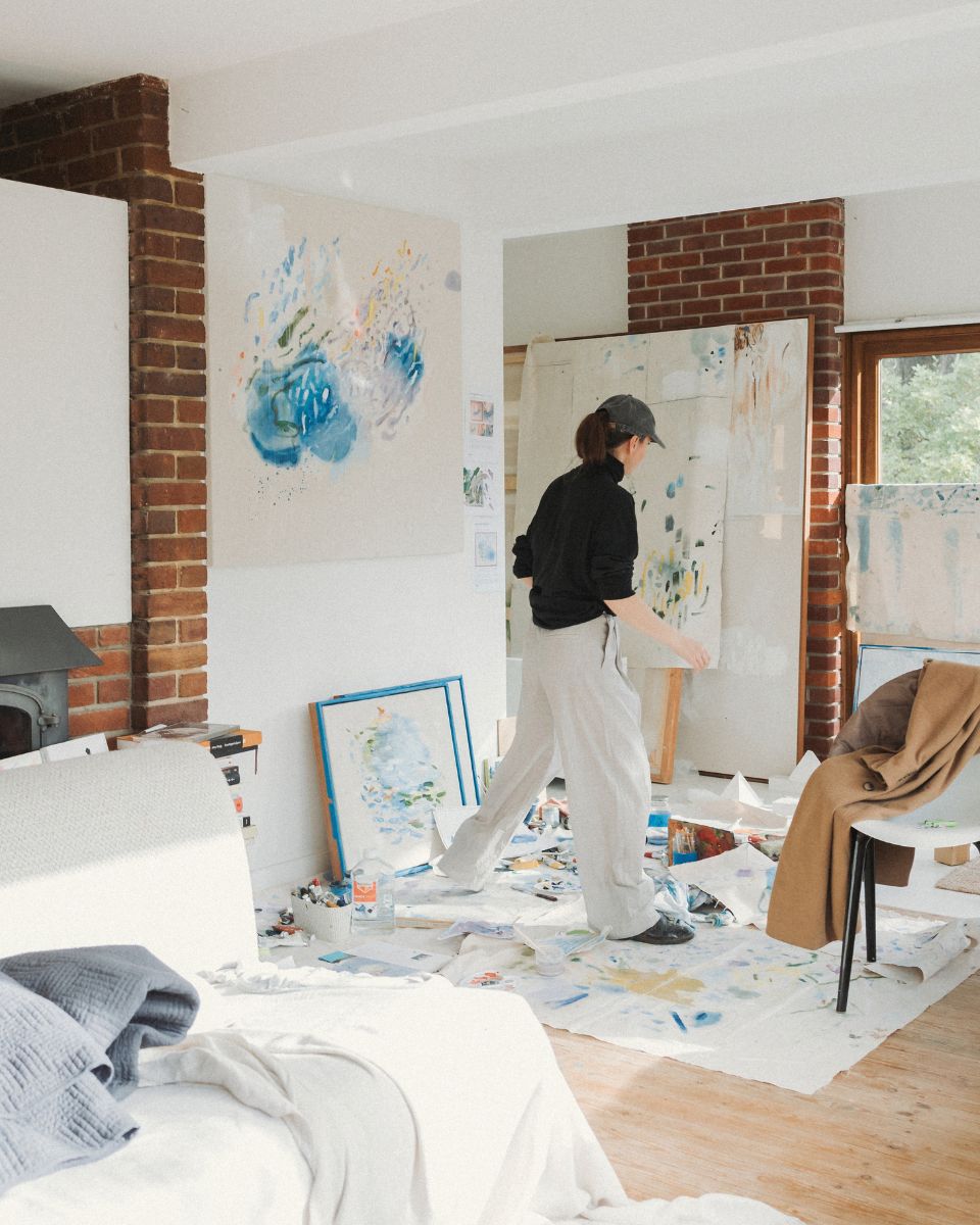 Artist Ita Wooller walking across her sunlit studio filled with abstract paintings, brushes, and drop cloths.