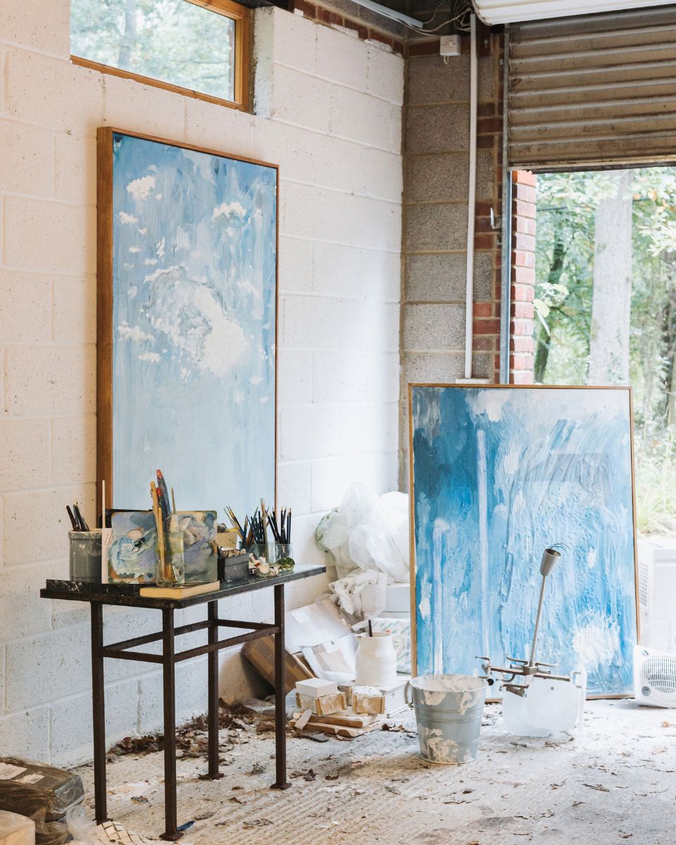 Large framed blue and white abstract oil paintings displayed against white brick walls near studio tools and window.
