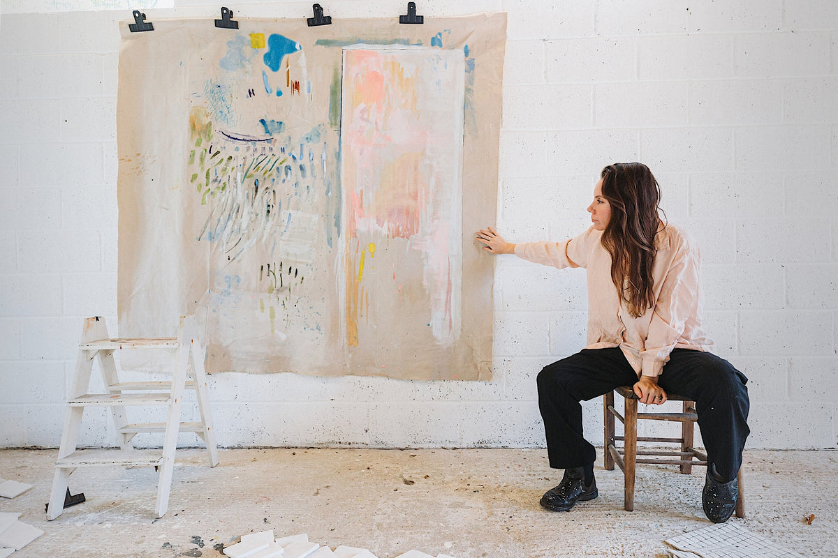Artist Ita Wooller seated on a stool beside a large un-stretched canvas painting mounted on a studio brick wall.
