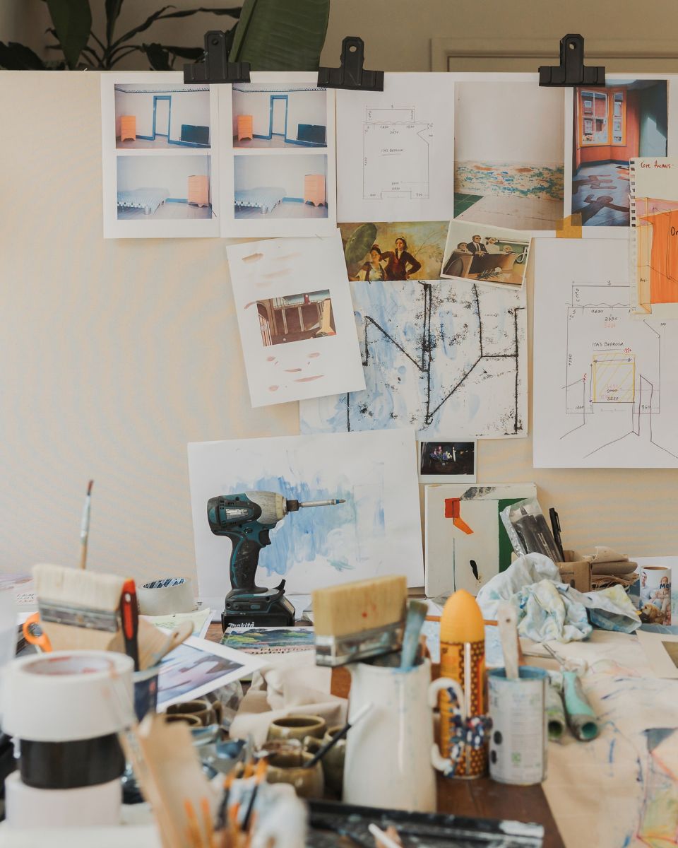 Studio workspace desk with paint jars, drill, and brushes in front of a wall pinned with architectural sketches and reference photos.