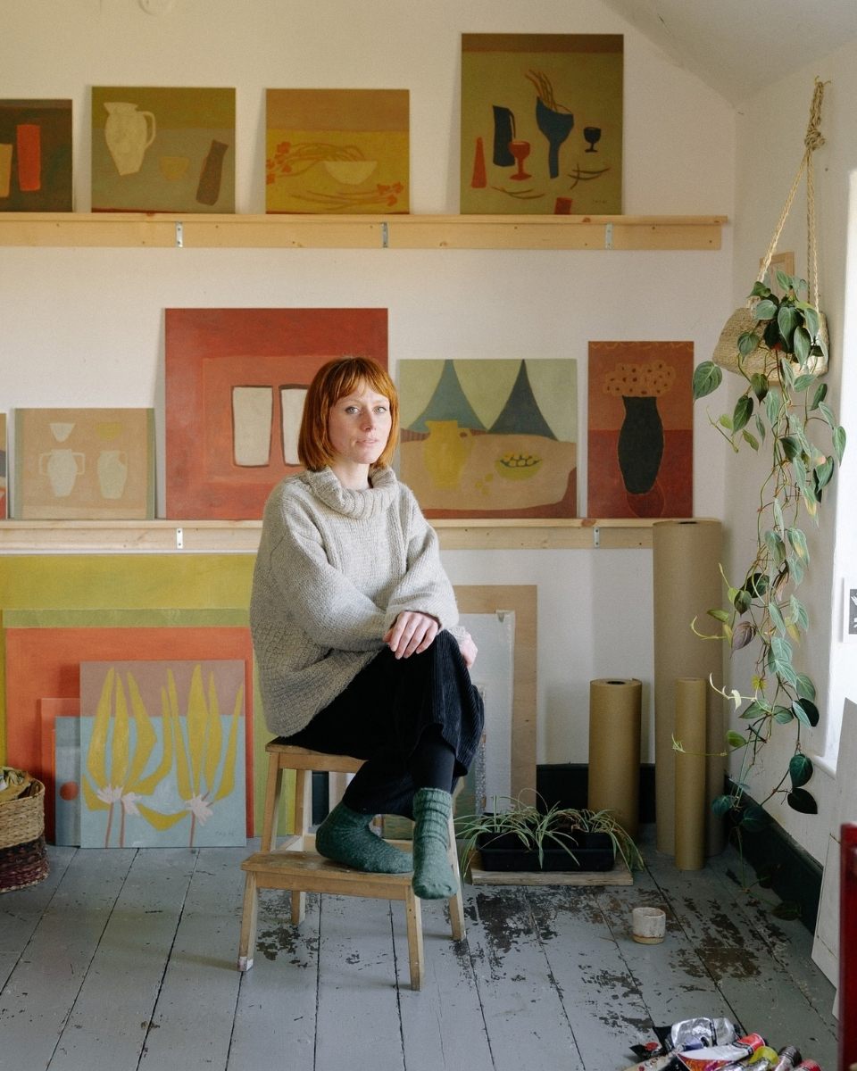 A seated portrait of artist Jay Harper in her home studio in front of her finished muted oil paintings.