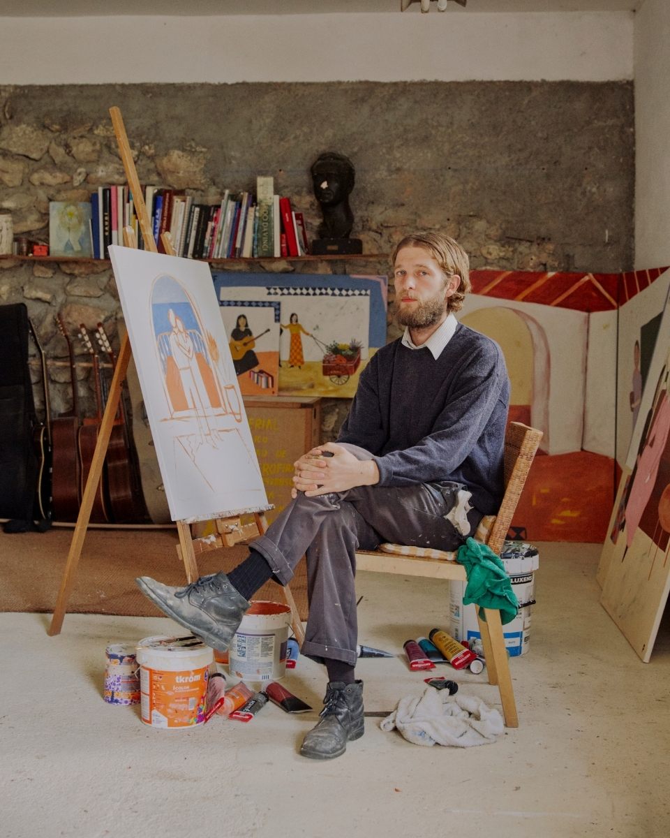 Artist Joshua Perkin sitting in his studio next to a work-in-progress figurative painting on an easel.