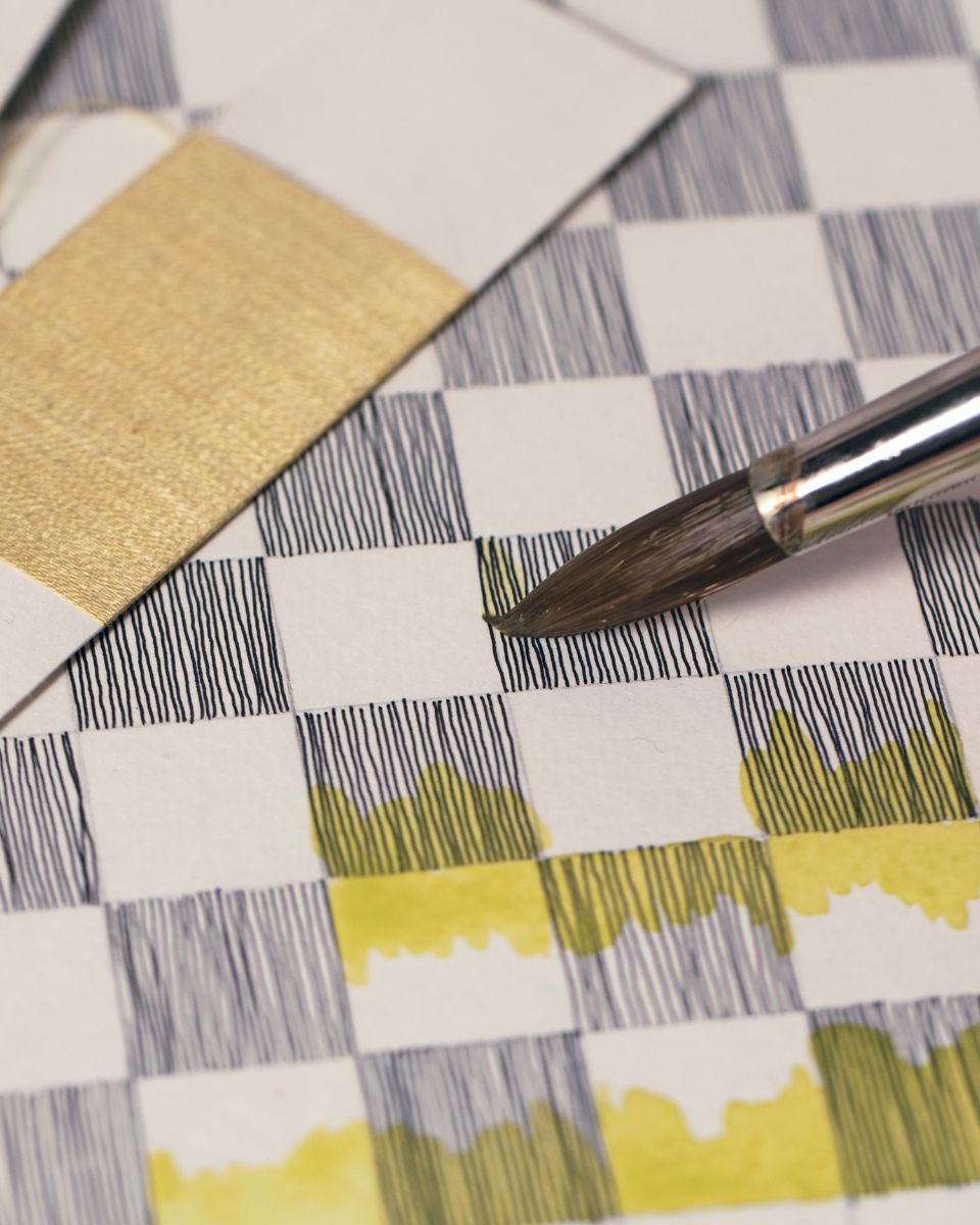 Fine detail of a paintbrush applying yellow watercolour to a black inked grid pattern study on paper.