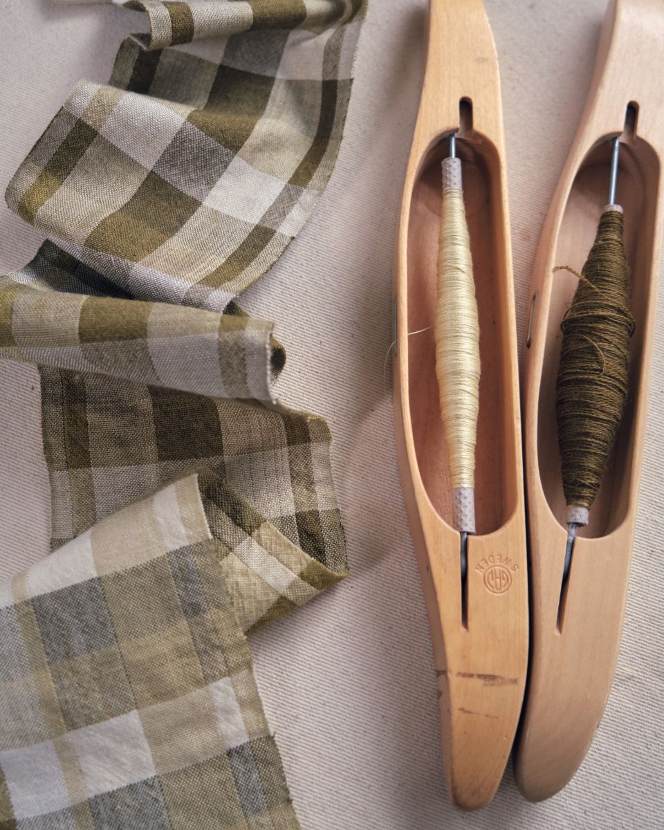 Two wooden weaving shuttles filled with neutral thread resting next to a folded olive-and-cream checkered woven textile.