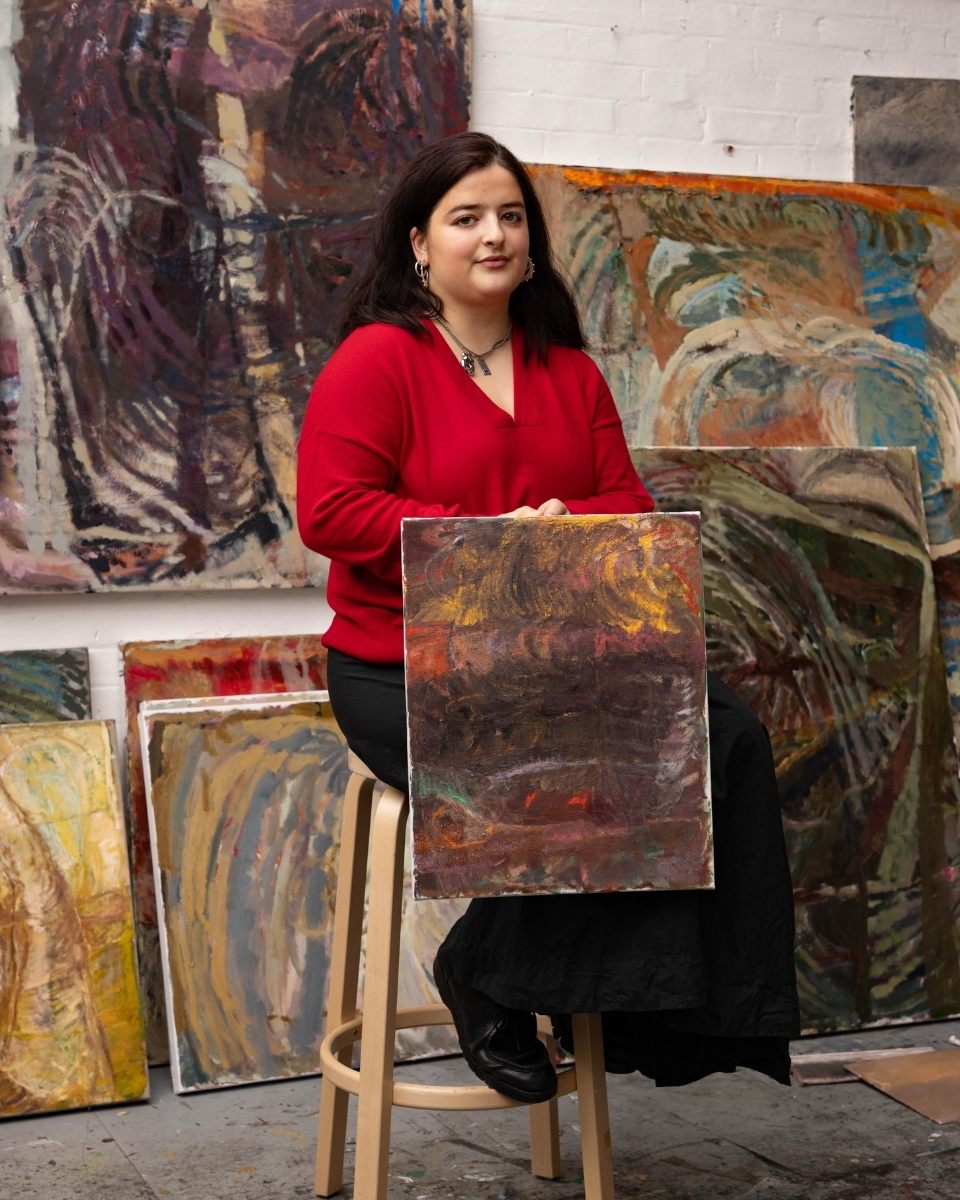 Portrait of Katarina Lalic in her studio, seated on a stool and holding an original oil painting amidst her larger abstract landscapes.