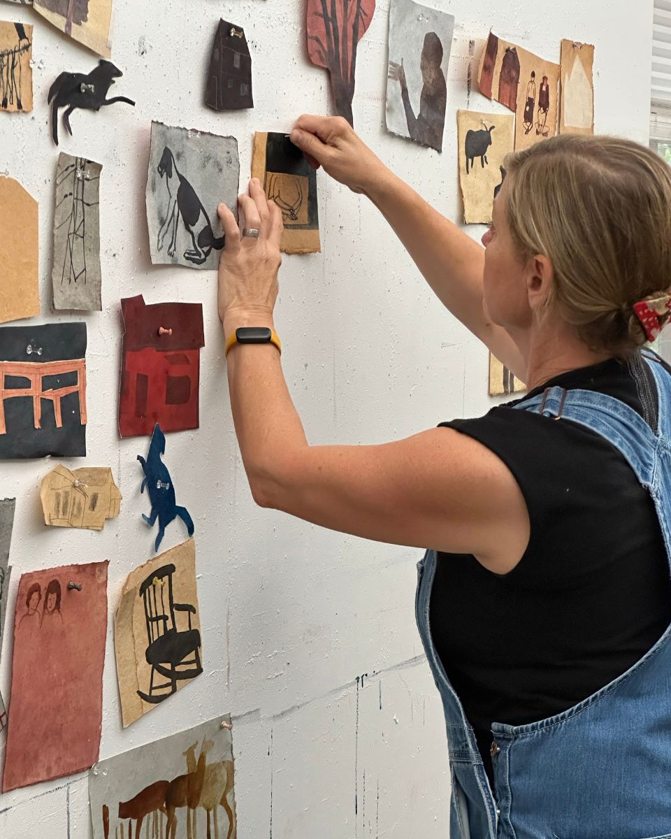Artist Kelly Popoff pinning small paper drawings and animal sketches onto a white studio wall grid display.