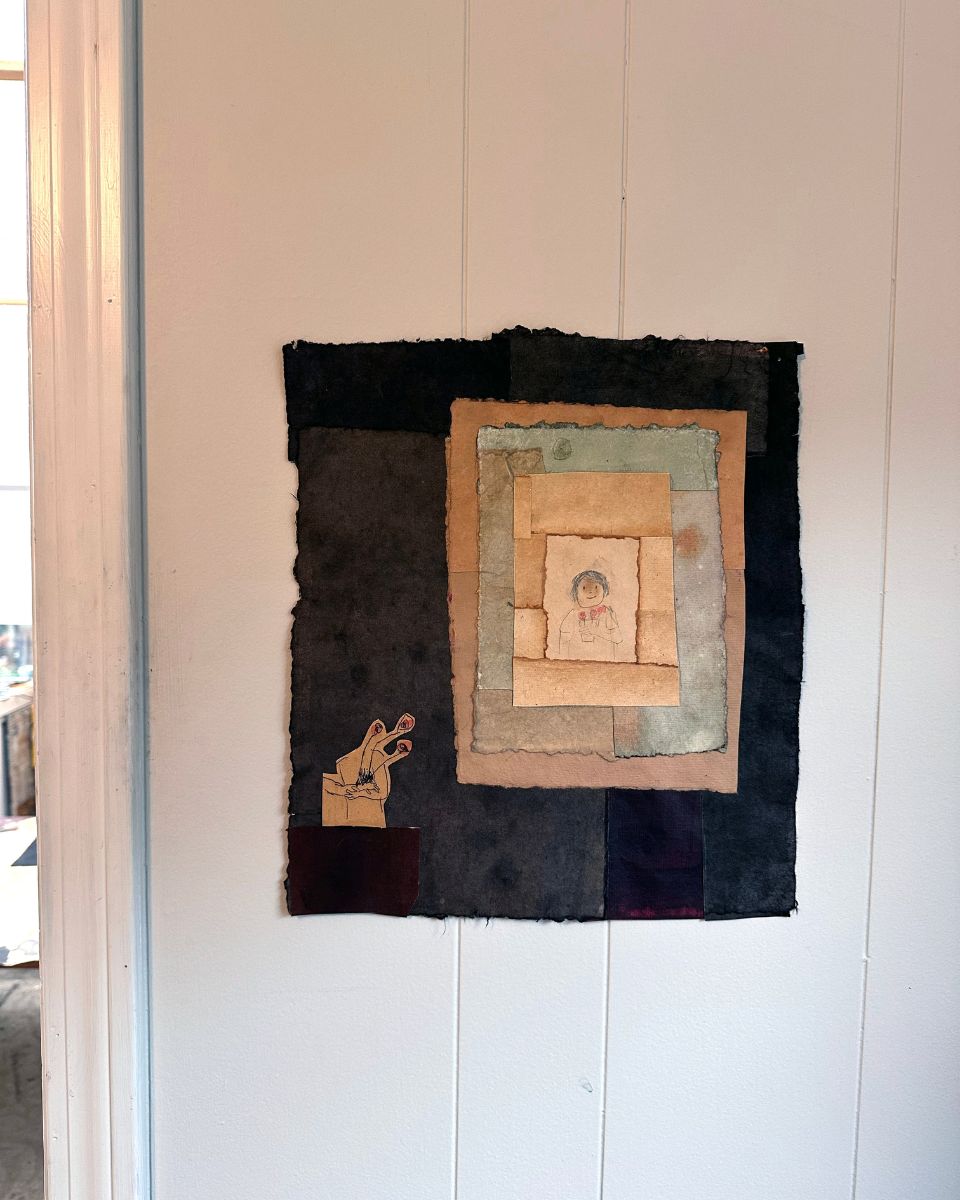 Layered mixed media paper collage depicting a central figure drawing mounted against a dark backdrop on a white wall.