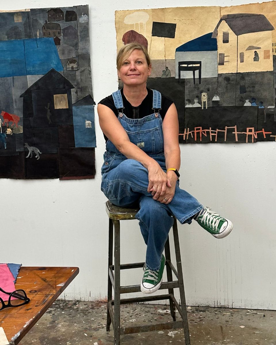 Artist Kelly Popoff sitting on a stool in her studio in denim dungarees with large quilt-style paintings displayed behind her.