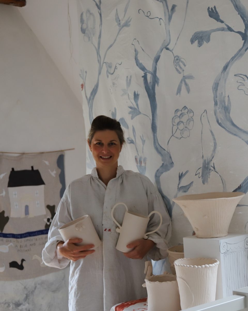 A smiling portrait of ceramicist Lisa Sandner holding two finished, hand-painted vases in her atelier.