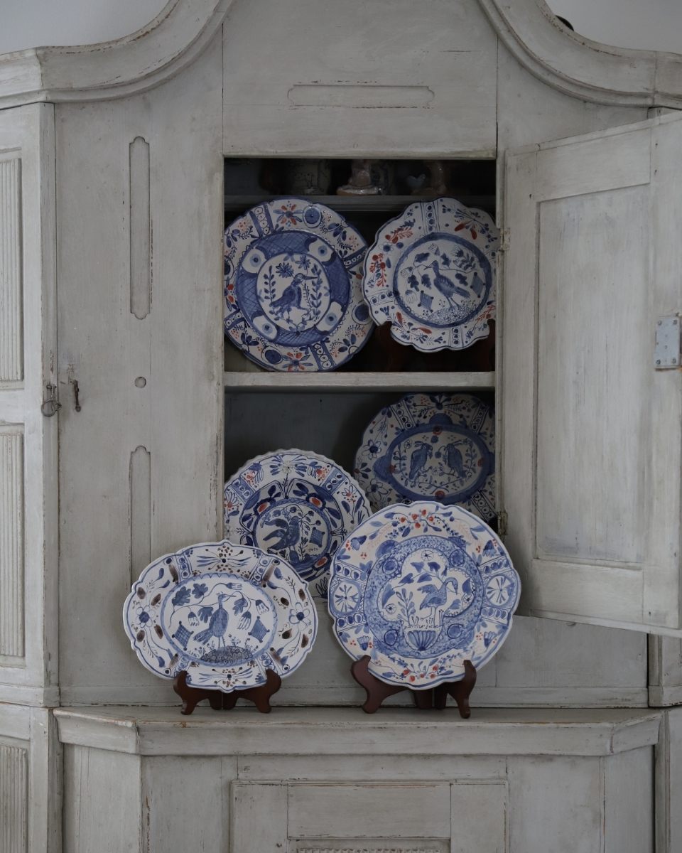 Lisa Sandner's hand-painted ceramic plates displayed inside an open light grey-washed wooden cabinet.